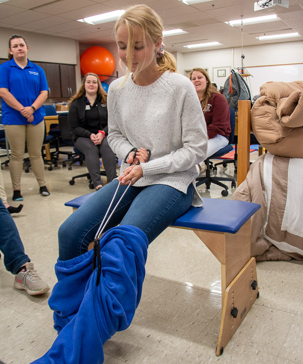 A high-schooler uses an adaptive tool to don a pair of sweatpants in the occupational therapy assistant lab.