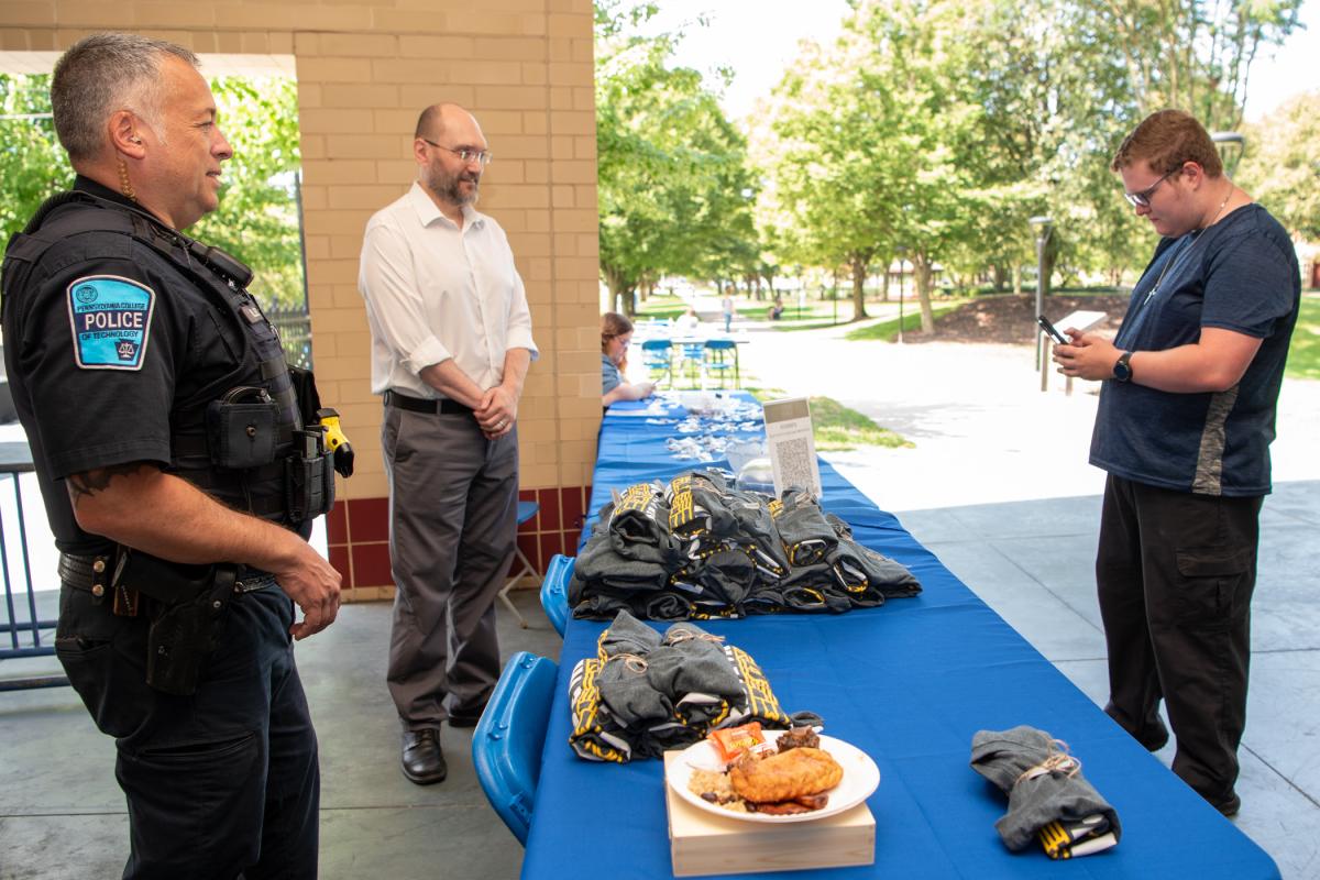 Electrical technology freshman Gavin A. Walsh registers his attendance with his cellphone and a QR code visual as “table staff” Penn College Police Chief David C. Pletz and Jeff Brown, vice president for information technology/chief information officer, greet attendees. (The plate in the foreground belongs to a student who, in yet more confirmation of campus security, asked Pletz to guard it while she ran an errand.)