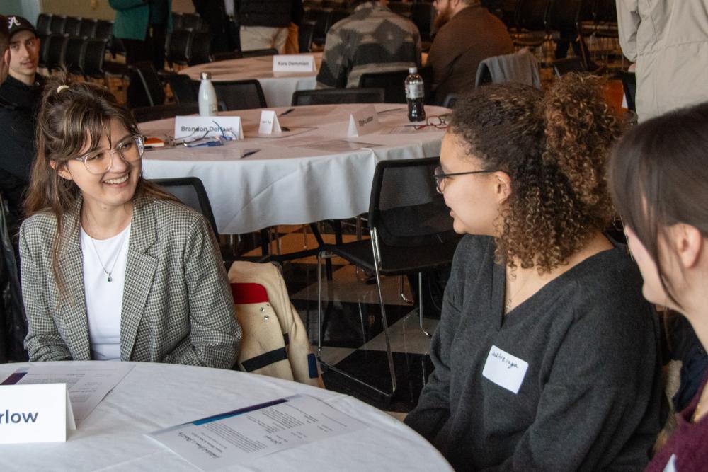 A graduate chats with students at a lunch table.