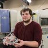 Joel Bergerstock caption: Penn College manufacturing engineering technology student Joel E. Bergerstock, of Liverpool, proudly displays one of three emblems he recently made for The Bicycle Center’s Susquehanna River Walk initiative. Bergerstock is standing in front of the electrical discharge machine he used to cut aluminum to form the emblems.